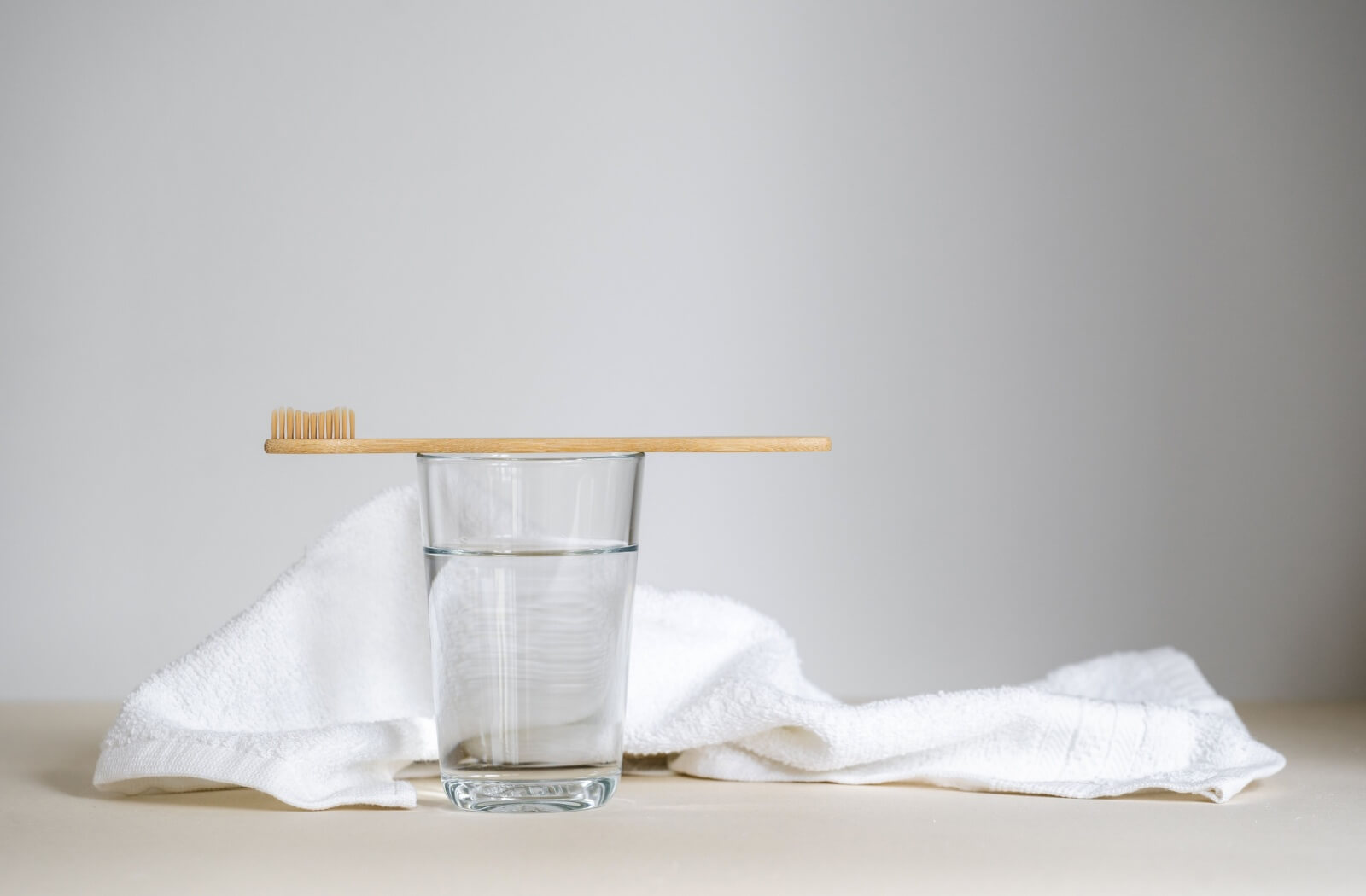 A picture of a clear glass of water with a toothbrush laying on top of the rim of the glass and a towel laying next to it.