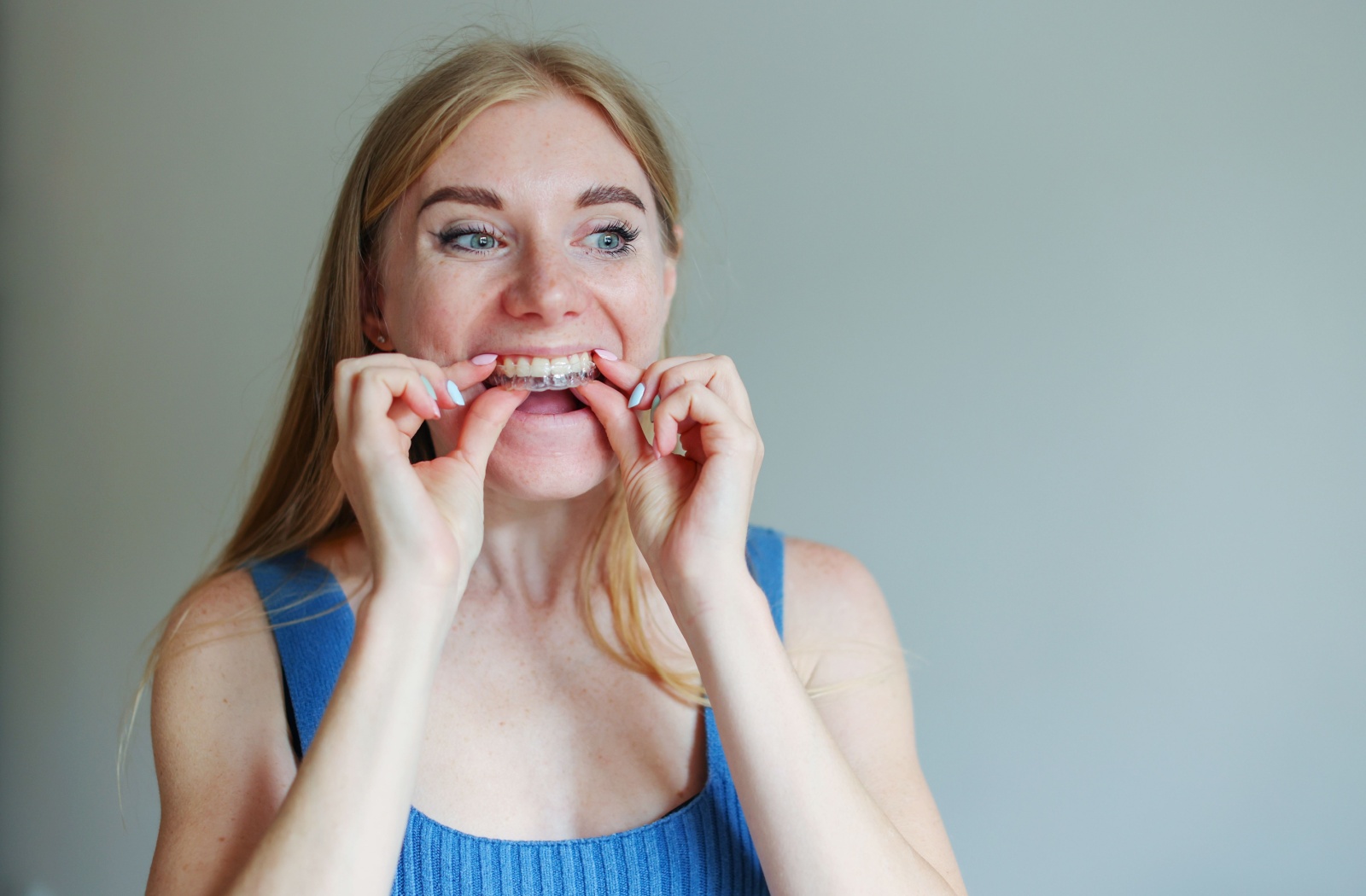 Person with long blond hair placing clear aligners on their teeth while smiling and looking in the mirror