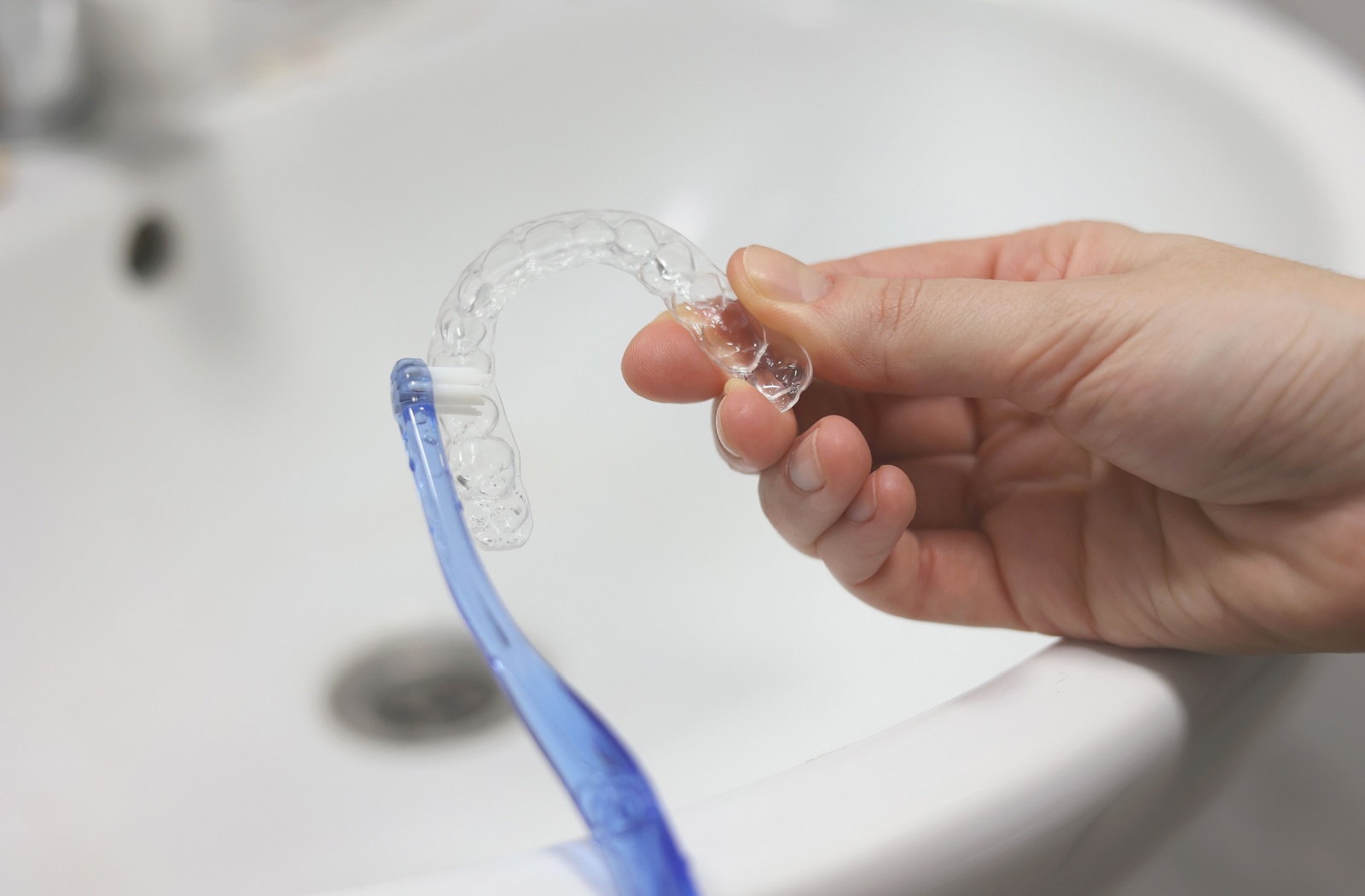 Hand holding clear aligner near bathroom sink with toothbrush gently cleaning the inside surface