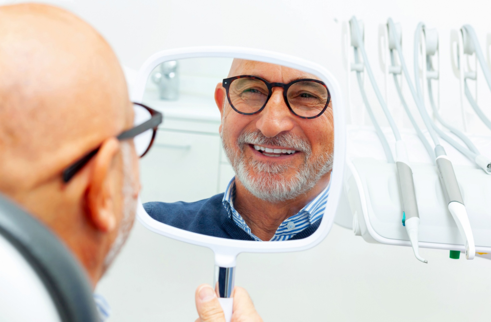 Older adult with glasses smiling in dental chair while examining dental implants in handheld mirror